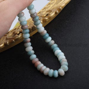 Pu&ograve; includere: Una collana di perline di pietre preziose nei toni del blu, bianco e pesca. Le perline rotonde sono infilate insieme, creando una collana o un braccialetto. Le perline sono presentate davanti a una cornice decorativa dorata.
