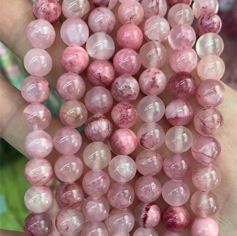 Puede incluir: Un collar de cuentas de piedras preciosas rosas. Las cuentas son redondas, con diferentes tonos de rosa y algunas con inclusiones rosa-rojo m&aacute;s oscuras. Las cuentas est&aacute;n ensartadas, listas para la fabricaci&oacute;n de joyas. El efecto general es delicado y femenino.