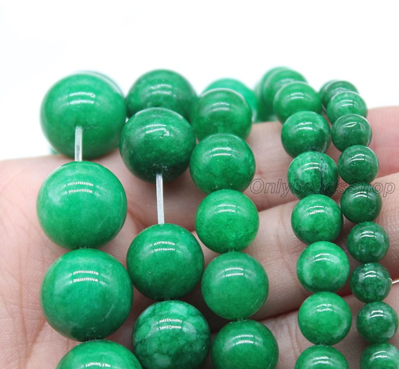 May include: A strand of round, green jade beads. The beads are smooth and polished, with a slight variation in size. The beads are strung on a clear elastic cord.