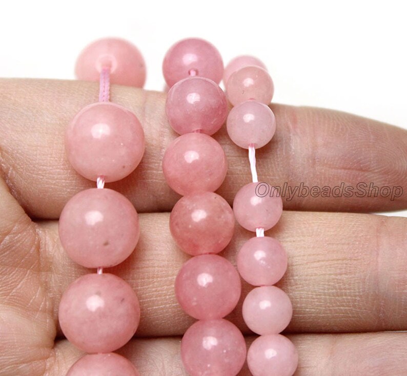 May include: A strand of pink rose quartz beads, with a variety of sizes, strung on a white cord. The beads are smooth and polished, with a subtle shimmer.