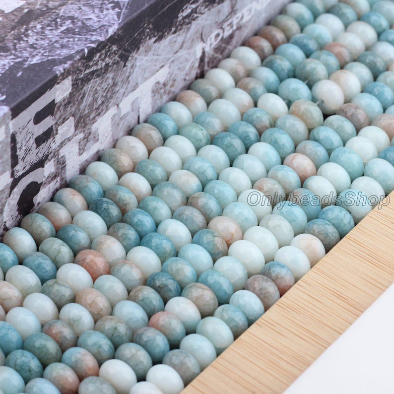 Pu&ograve; includere: Primo piano di una scatola di legno piena di file di perline ovali lucide. Le perline sono un mix di blu pallido, bianco e pesca. La scatola ha un disegno stampato sul lato. Il testo "OnlybeadsShop" &egrave; visibile.