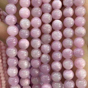 May include: A close-up of strands of light pink, round gemstone beads. The beads have a smooth, polished surface and a subtle, pearlescent sheen. The beads are uniform in size and shape, suggesting they are ready for jewelry making.