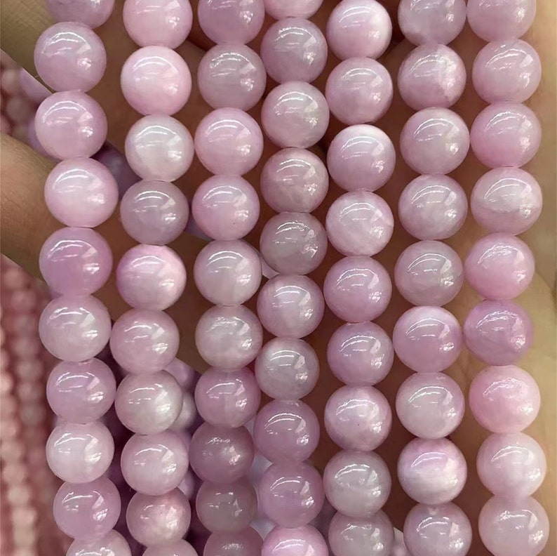 Puede incluir: Primer plano de hilos de cuentas de piedras preciosas redondas de color rosa claro. Las cuentas tienen una superficie lisa y pulida y un brillo sutil y nacarado. Las cuentas son uniformes en tama&ntilde;o y forma, perfectas para la fabricaci&oacute;n de joyas.