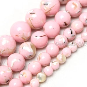 Pode incluir: Um fio de contas de concha rosa e branca com detalhes marrons e dourados. As contas são redondas e têm um acabamento brilhante. As contas são enfiadas em um fio transparente. As contas têm aproximadamente 10 mm de diâmetro.