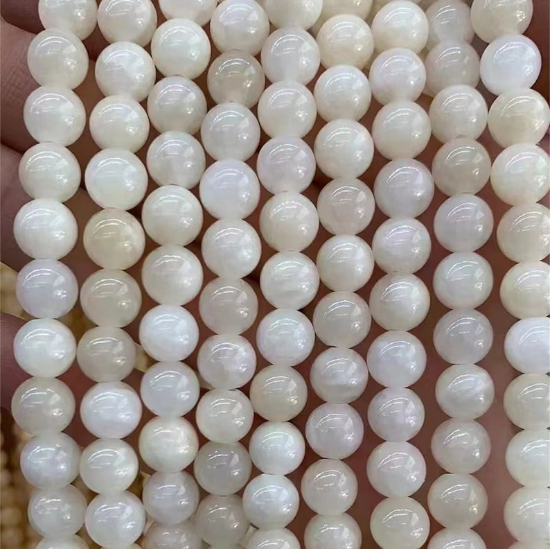 Puede incluir: Hilo de cuentas blancas redondas y luminosas. Las cuentas tienen un brillo nacarado, que refleja la luz. Las cuentas est&aacute;n ensartadas, lo que sugiere que son para la fabricaci&oacute;n de joyas o manualidades. La paleta de colores general es suave y neutra.