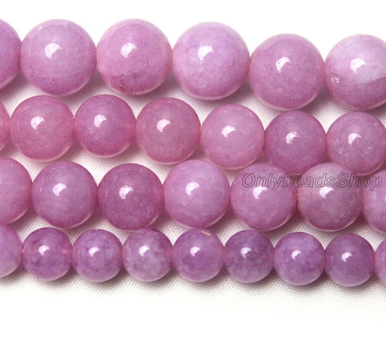 Puede incluir: Una hebra de cuentas redondas de piedras preciosas de color lavanda. Las cuentas son lisas y pulidas, con un ligero brillo. Las cuentas est&aacute;n dispuestas en un patr&oacute;n escalonado, con las cuentas m&aacute;s grandes en el centro y las m&aacute;s peque&ntilde;as en los bordes.