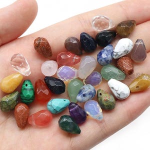 Può includere: Una collezione di perline di pietre preziose a forma di goccia in vari colori, tra cui trasparente, giallo, verde, rosso, blu, viola e marrone. Le perline sono sfaccettate e lisce, con finiture opache e traslucide.