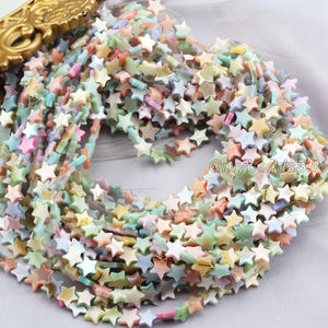 Puede incluir: Hebras de cuentas en forma de estrella de colores pastel, incluyendo rosa, verde y naranja. Las cuentas están ensartadas, creando una exhibición texturizada y colorida. Las cuentas son probablemente para la fabricación de joyas.