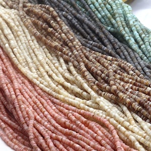 Könnte beinhalten: Nahaufnahme von mehreren Strängen kleiner, flacher Muschelkernperlen in verschiedenen Farben. Die Farben umfassen Schattierungen von Rosa, Creme, Braun, Grau und Türkis. Die Perlen sind aufgefädelt und erzeugen eine strukturierte und farbenfrohe Darstellung.