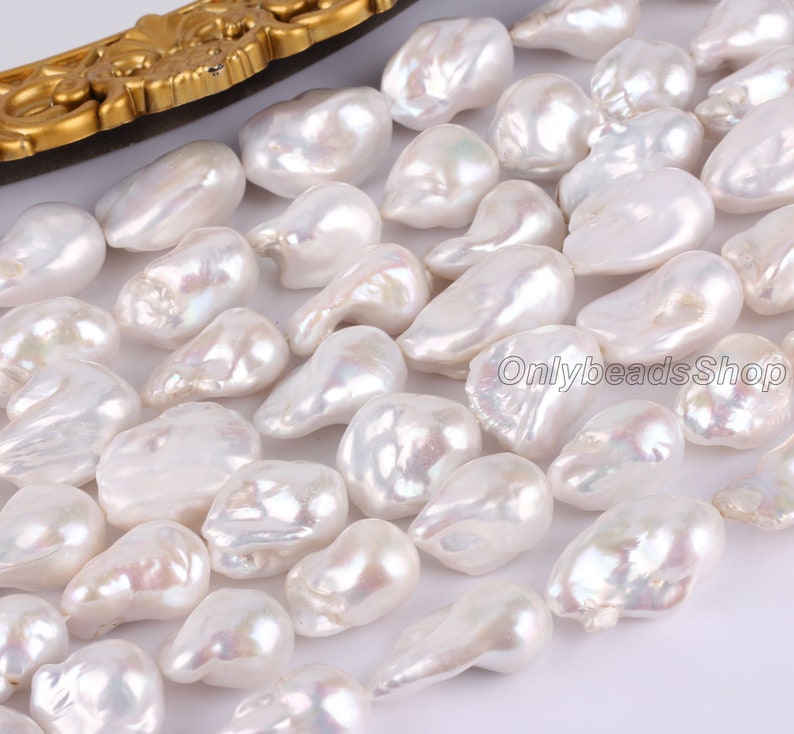 Peut inclure: Un brin de perles baroques blanches, avec un &eacute;clat iridescent. Les perles sont de forme et de taille irr&eacute;guli&egrave;res, et sont enfil&eacute;es sur un fil transparent. Les perles sont pr&eacute;sent&eacute;es sur un fond blanc.