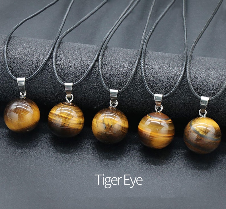May include: Five necklaces with black cords and round tiger's eye stone pendants. The pendants are all the same size and shape, and they have a golden brown color with a slight shimmer.