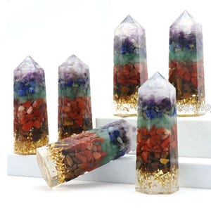 May include: Six clear crystal chakra wands with seven layers of different colored gemstones, including amethyst, lapis lazuli, green aventurine, carnelian, tiger's eye, and clear quartz. The wands are topped with a clear quartz point and have gold leaf accents.