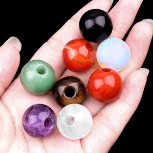 May include: A collection of gemstone beads in various colors, including green, red, black, and purple. Each bead is round with a hole drilled through the center, suitable for stringing. The beads are displayed against a dark background.