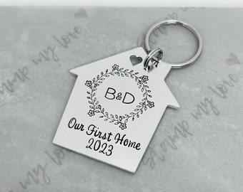 May include: Silver house-shaped keychain with a heart cut-out and a floral wreath with the initials "B&D" and the text "Our First Home 2023".