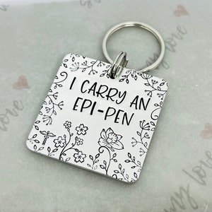 May include: A silver keychain with a square tag that says "I carry an epi-pen" with a floral design.
