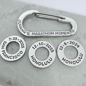 May include: Four silver metal keychains with engraved text. The keychains are in the shape of a circle and a carabiner. The text on the keychains reads "7-23-2023 SAN FRANCISCO", "12-10-2023 HONOLULU", "12-8-2024 HONOLULU", and "JUHANG'S MARATHON MOMENTS".