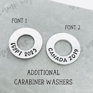 May include: Two silver metal round washers with a hole in the centre. The washer on the left has the text "EGYPT 2023" and the washer on the right has the text "CANADA 2019". The text is in black. The washers are on a white surface. The text "FONT 1" is above the left washer and the text "FONT 2" is above the right washer. The text "ADDITIONAL CARABINER WASHERS" is below the washers.