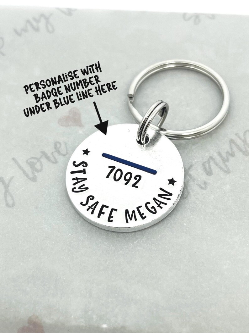 May include: A silver round keychain tag with the text "STAY SAFE MEGAN" and the badge number "7092" above a blue line. The text "PERSONALISE WITH BADGE NUMBER UNDER BLUE LINE HERE" is also visible.