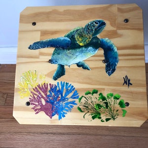May include: A wooden square with a painted sea turtle swimming above coral. The coral is painted in shades of yellow, red, blue, and green. The turtle is painted in shades of blue and green. The background is a light wood color. The text "HK" is painted in black in the bottom right corner.