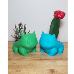 May include: Two colorful Bulbasaur figurines, one green and one blue, sit on a wooden surface. Behind them are small potted plants, including a zebra plant and a cactus with a red top. The figurines are detailed and stylized.