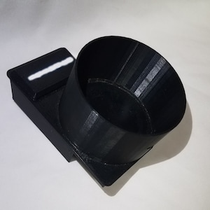 May include: A black 3D printed cup holder with a small rectangular compartment on the side. The cup holder is designed to fit a standard-sized cup.