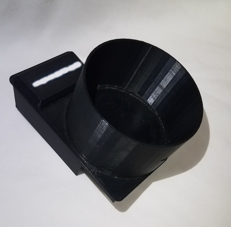 May include: A black 3D printed cup holder with a small rectangular compartment on the side. The cup holder is designed to fit a standard-sized cup.