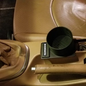 May include: A close-up of a car's interior with a brown leather center console. The console has a cup holder, a gear shifter, and a handbrake. The cup holder is a black plastic cup holder with the word "Porsche" printed on it.