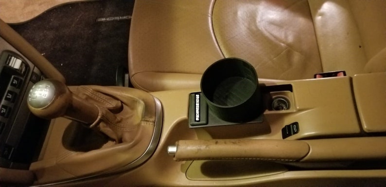 May include: A close-up of a car's interior with a brown leather center console. The console has a cup holder, a gear shifter, and a handbrake. The cup holder is a black plastic cup holder with the word "Porsche" printed on it.