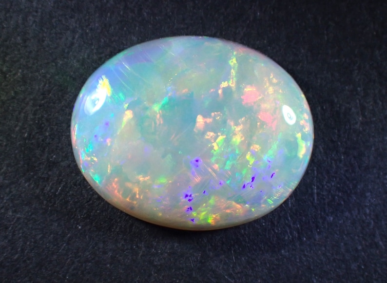May include: A white opal gemstone with a light green, blue, and pink iridescent sheen. The opal is oval shaped and has a smooth, polished surface.