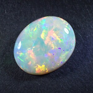 May include: A white opal gemstone with a light blue, green, and purple iridescent sheen. The oval-shaped stone is set against a dark grey background.