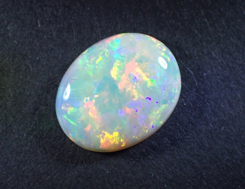 May include: A white opal gemstone with a light blue, green, and purple iridescent sheen. The oval-shaped stone is set against a dark grey background.