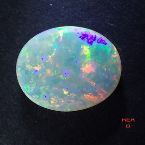 May include: A white opal gemstone with iridescent flashes of green, blue, and purple. The opal is oval shaped and has a smooth, polished surface.