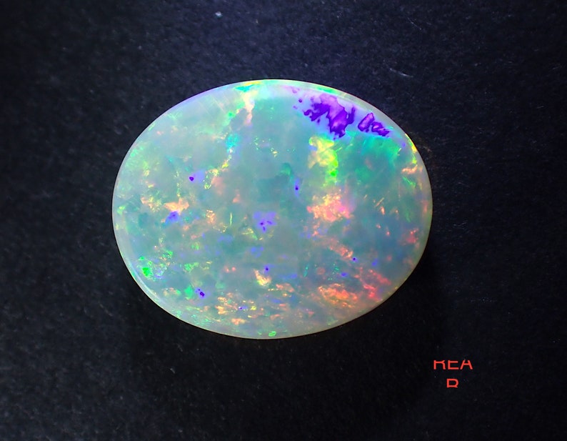 May include: A white opal gemstone with iridescent flashes of green, blue, and purple. The opal is oval shaped and has a smooth, polished surface.