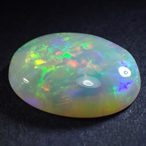 May include: A white opal gemstone with green, blue, and red flashes of colour. The oval-shaped stone is set against a black background.