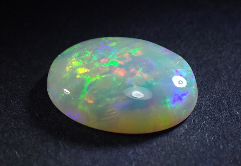 May include: A white opal gemstone with green, blue, and red flashes of colour. The oval-shaped stone is set against a black background.