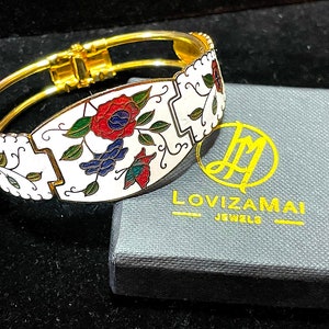 Puede incluir: Un brazalete de oro con un diseño de esmalte blanco con motivos florales rojos, azules y verdes. El brazalete está en una caja gris con el texto "Lovizamai Jewels".