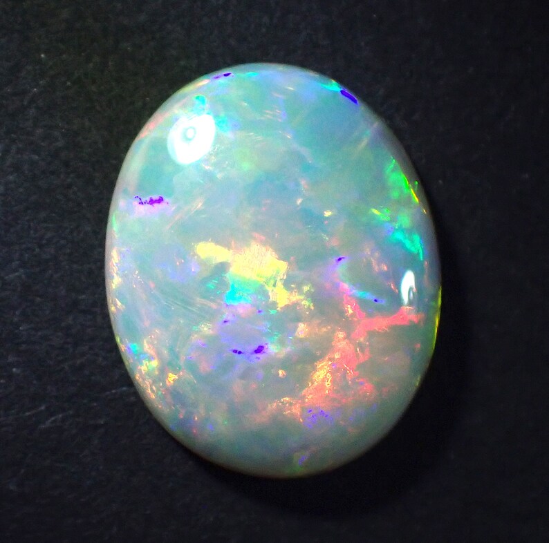May include: A white opal gemstone with a light blue, green, and pink iridescent sheen. The opal is oval shaped and has a smooth, polished surface.