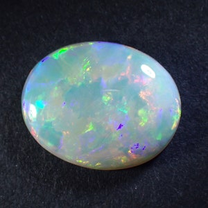 May include: A white opal gemstone with green, blue, and purple iridescent flashes. The oval-shaped stone is set against a black background.