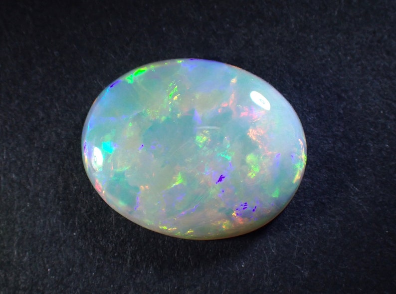May include: A white opal gemstone with green, blue, and purple iridescent flashes. The oval-shaped stone is set against a black background.