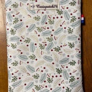 Pode incluir: Uma bolsa branca com zíper com um padrão repetido de folhas verdes e douradas, bagas vermelhas e pequenos pontos brancos. A bolsa tem um zíper vermelho e uma etiqueta que diz "Croixpatch74". Uma pequena bandeira francesa está presa na lateral.