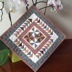 May include: A square quilted wall hanging with a geometric design. The quilt features a central diamond pattern in shades of brown, white, and gray, surrounded by borders of gray, white, and pink. The quilt is approximately 12 inches square.
