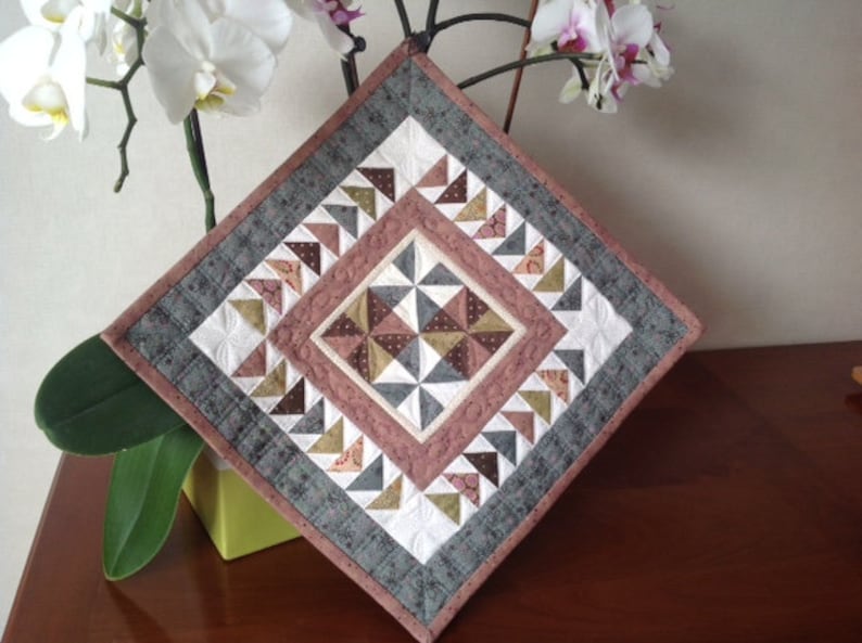 May include: A square quilted wall hanging with a geometric design. The quilt features a central diamond pattern in shades of brown, white, and gray, surrounded by borders of gray, white, and pink. The quilt is approximately 12 inches square.