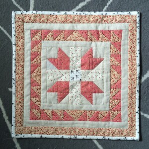 May include: A square quilted wall hanging with a central star design in pink and white fabrics. The star is surrounded by a border of triangles and a patterned edge with a black and white trim. The design is symmetrical and handmade.