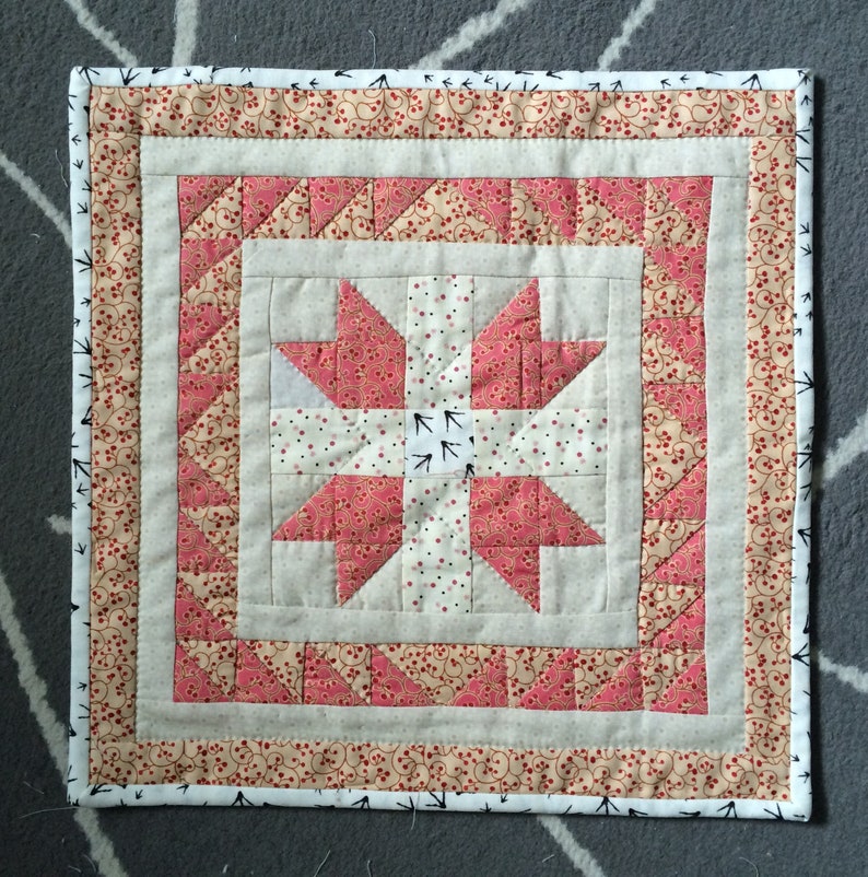 May include: A square quilted wall hanging with a central star design in pink and white fabrics. The star is surrounded by a border of triangles and a patterned edge with a black and white trim. The design is symmetrical and handmade.