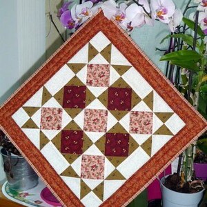 May include: Square quilted patchwork table topper with a geometric star and square pattern. The design features squares of floral fabric in shades of red and pink, surrounded by white, brown, and tan triangles, and a rust-coloured border.