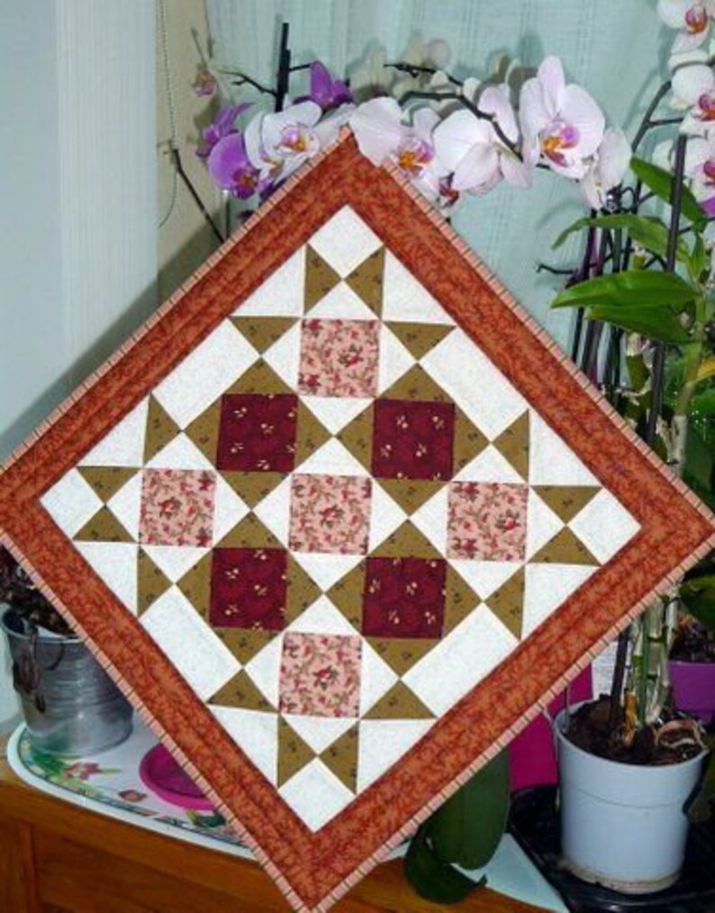 May include: Square quilted patchwork table topper with a geometric star and square pattern. The design features squares of floral fabric in shades of red and pink, surrounded by white, brown, and tan triangles, and a rust-coloured border.