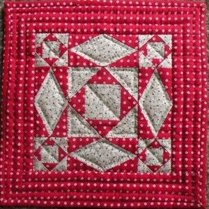May include: Square quilted potholder with a red polka dot border and a central geometric design in red and gray. The center features diamond and square shapes, creating a patchwork effect. The red border is also quilted.