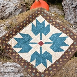 May include: A quilted, square-shaped table topper with a central design of teal blue stylized floral shapes on a cream background. The border is brown with a repeating leaf pattern. A small orange gourd rests in the background.