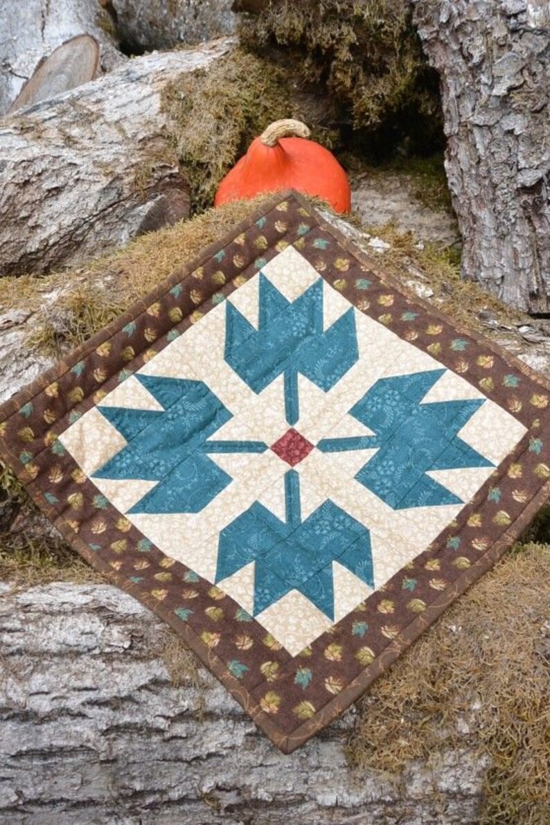 May include: A quilted, square table topper featuring a central design of teal blue stylized floral shapes on a cream background. The border is brown with a repeating leaf pattern. An orange gourd is in the background.