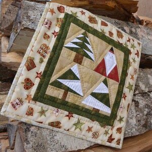 May include: A square Christmas-themed quilted wall hanging. The centre features a patchwork design of Christmas trees and Santa Claus. The border is cream-coloured with repeating patterns of stars, gifts, and hearts. The quilt is displayed on a stack of firewood.